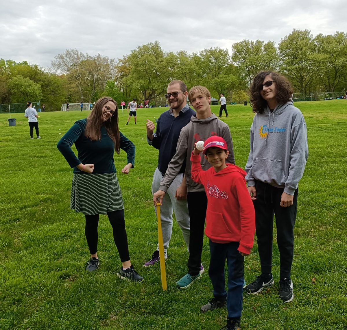 A few of our youth (and one of our leaders) strike poses in a grassy field with a whiffle ball and yellow bat.