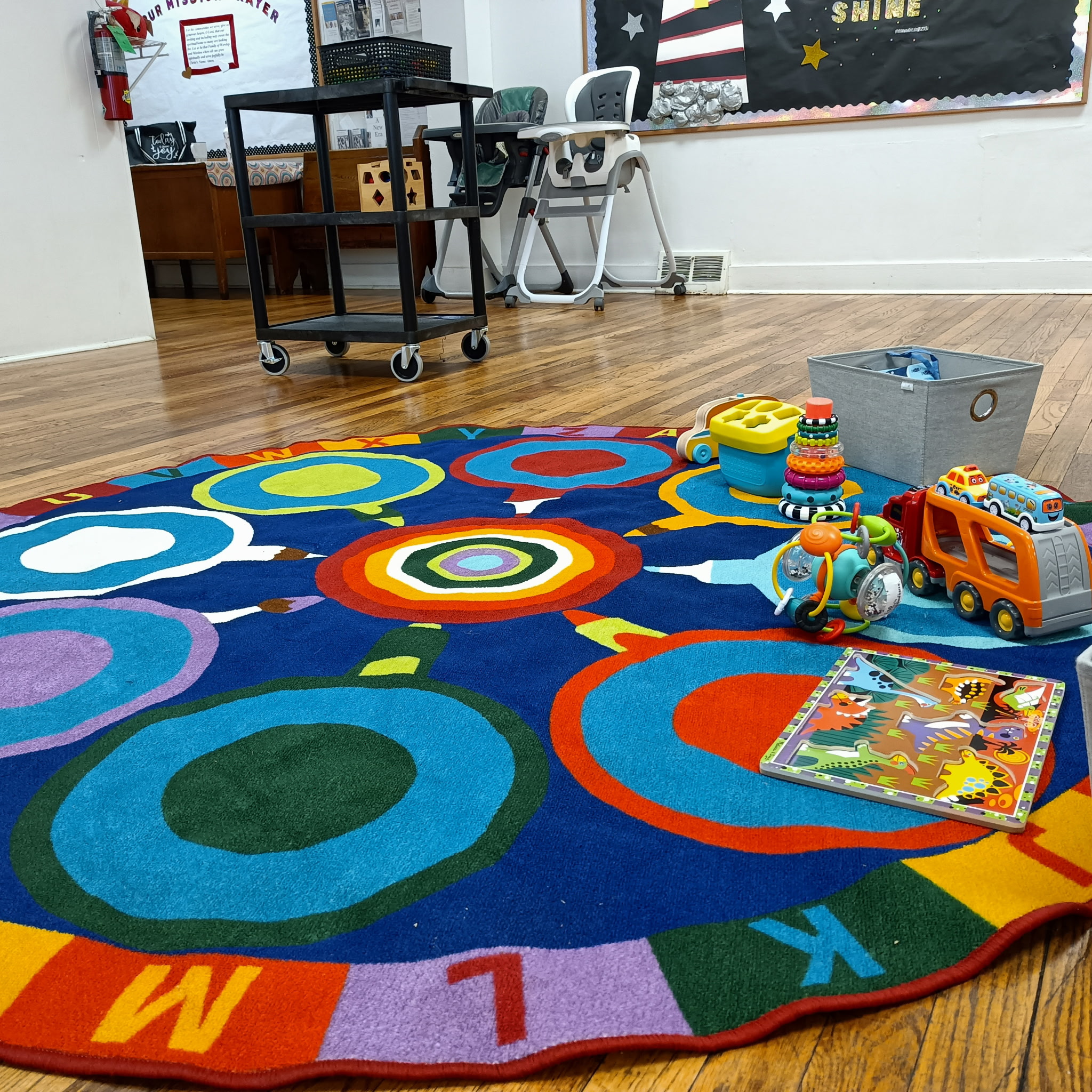 a colorful round rug with letters of the alphabet in colored blocks around the perimeter, with a few toys scattered on the right, a high chair and bulletin board visible in the background